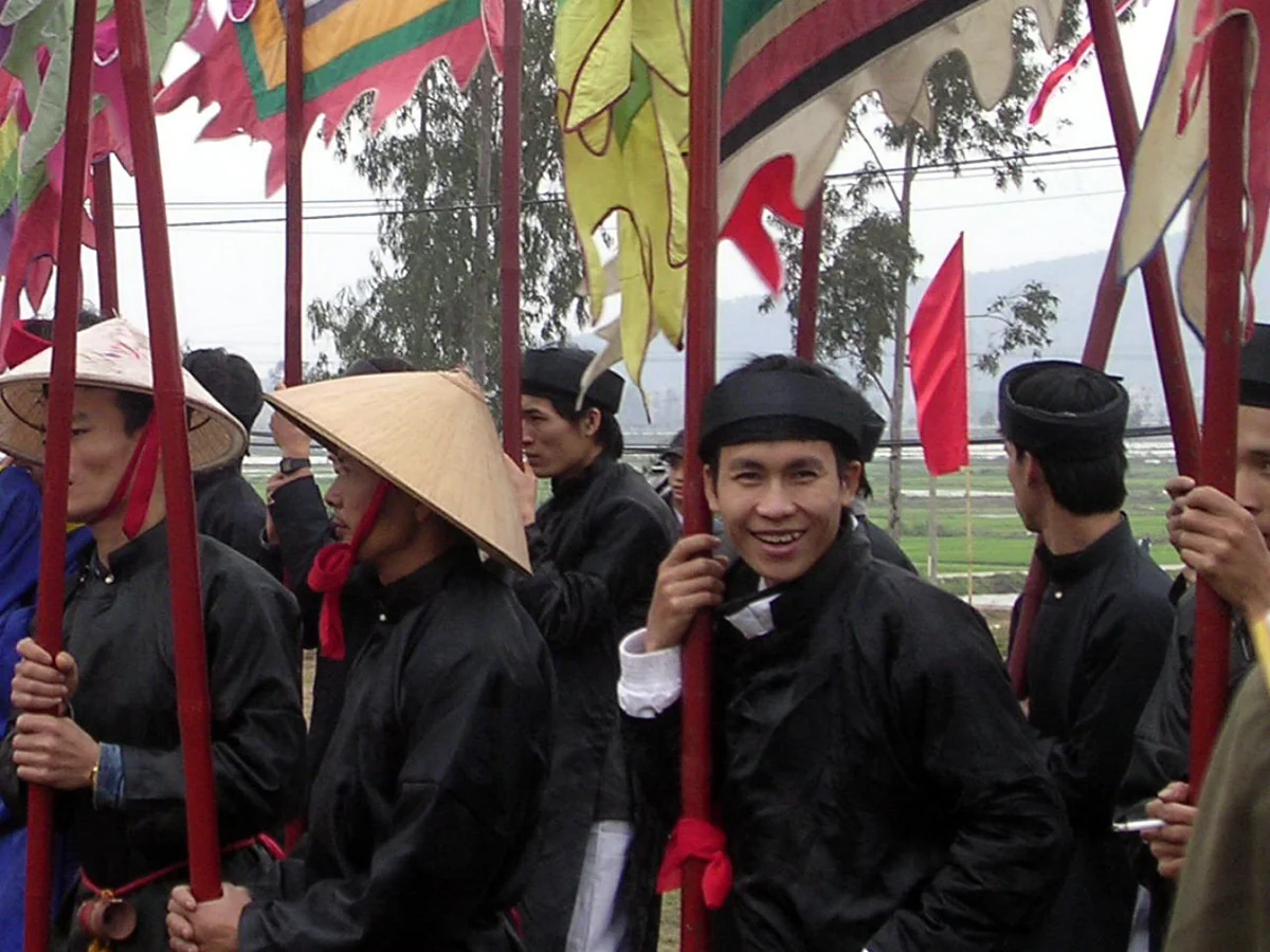 People in traditional attire hold colorful flags during a vibrant outdoor festival. A man smiles. Background shows trees and mountains.