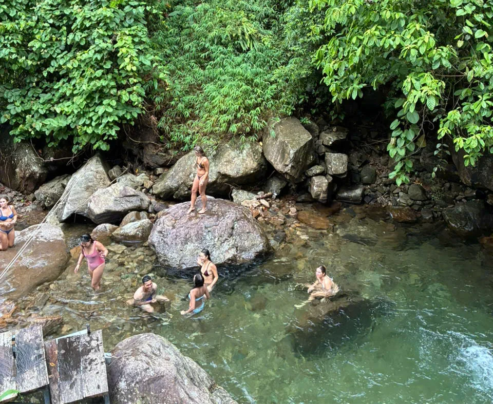 People swim and relax in a clear, rocky stream surrounded by lush green foliage. One person stands on a rock. The mood is joyful and serene.