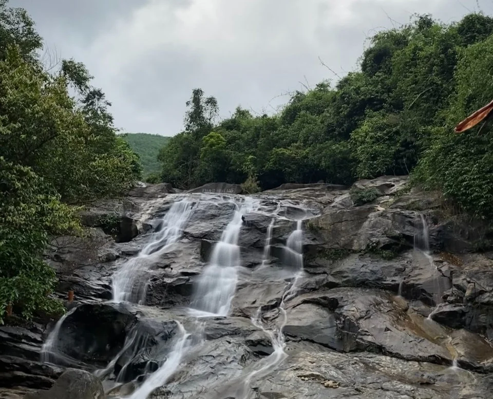 Cascading waterfall over smooth rocks, surrounded by lush green trees under a cloudy sky. Serene and natural setting.