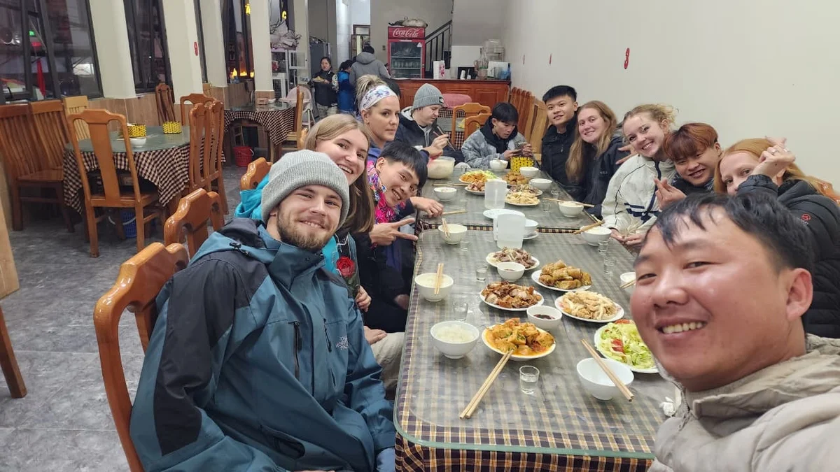Group of people smiling and dining in a cozy restaurant. Plates of food on the table. Wooden chairs and checkered tablecloths in the background.