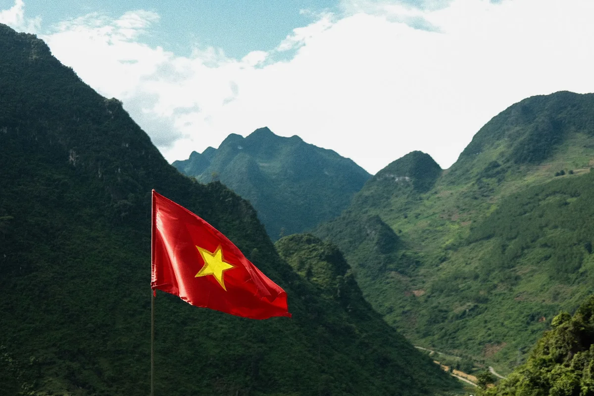 Vietnamese flag with a yellow star waves against a backdrop of lush green mountains and a cloudy sky, creating a serene and patriotic scene.