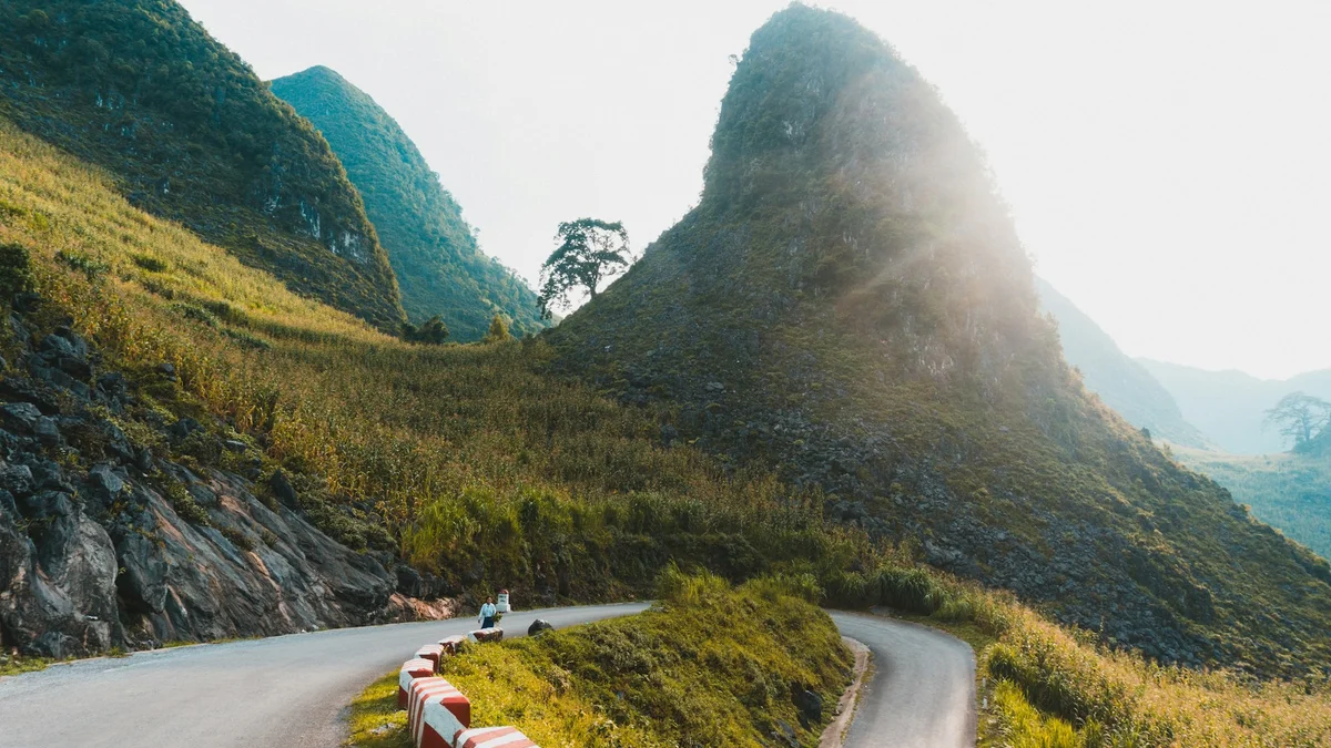 Winding road through lush green mountains with morning sunlight. Two people walk along the roadside, surrounded by vibrant greenery.