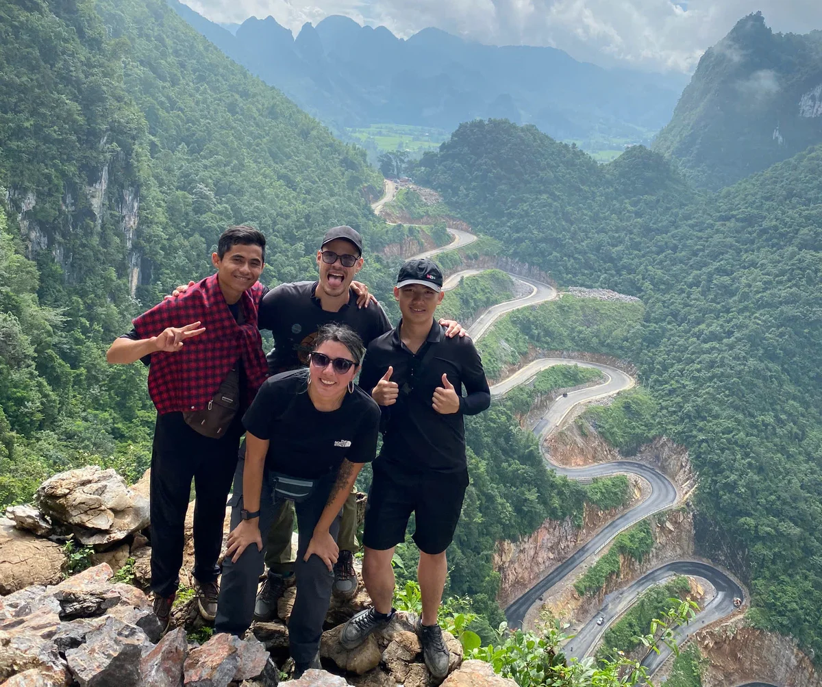 Four people pose happily on a rocky cliff with lush green mountains and winding roads in the background. Bright, overcast day.