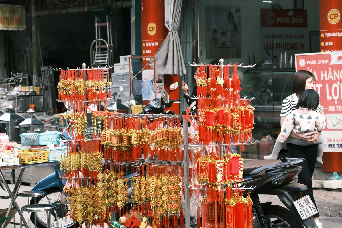 Street market with a display of gold and red Chinese decorations. A woman holds a child nearby. Background includes a pork shop and motorcycle.