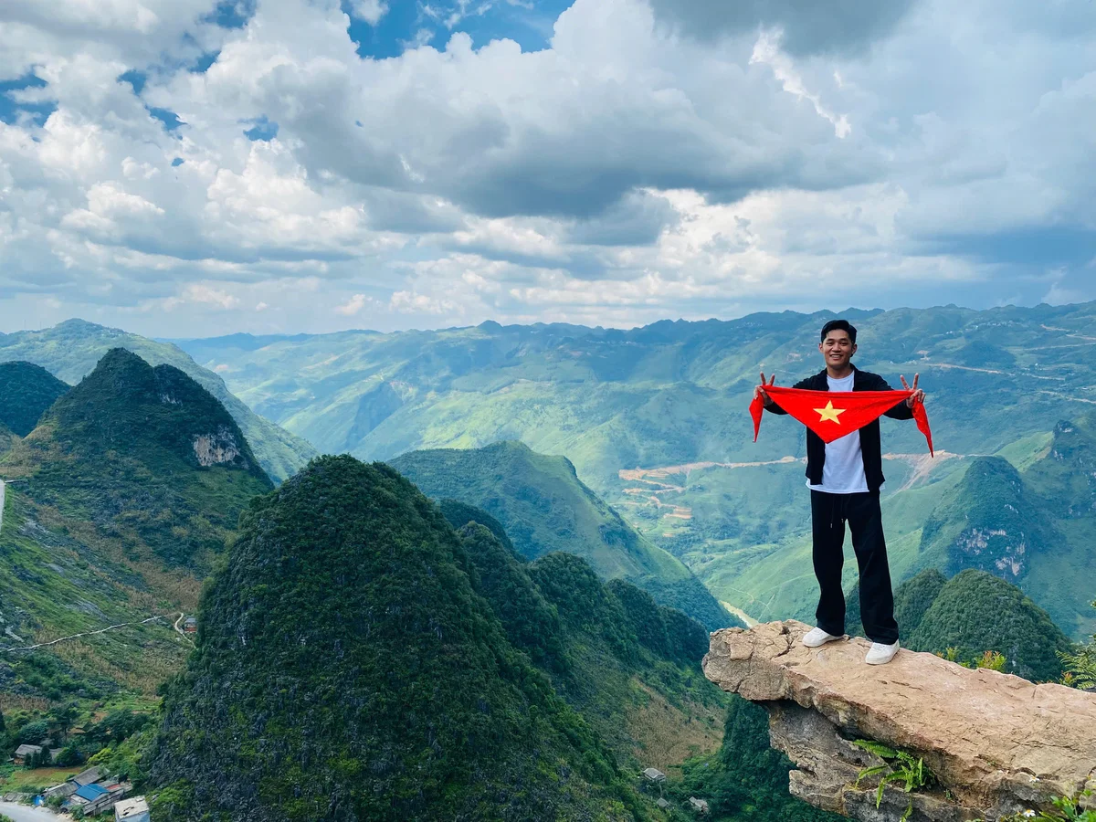 Man holding a Vietnamese flag on a cliff with lush green mountains in the background. The sky is partly cloudy, creating a serene mood.
