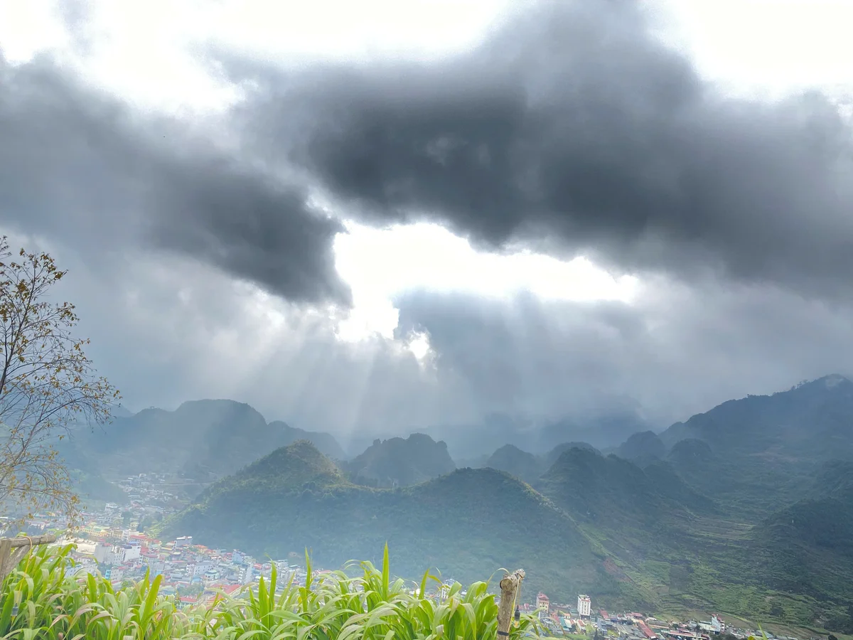Dramatic dark clouds with sunrays over a lush green mountain landscape and a small colorful town below, creating a serene, moody atmosphere.