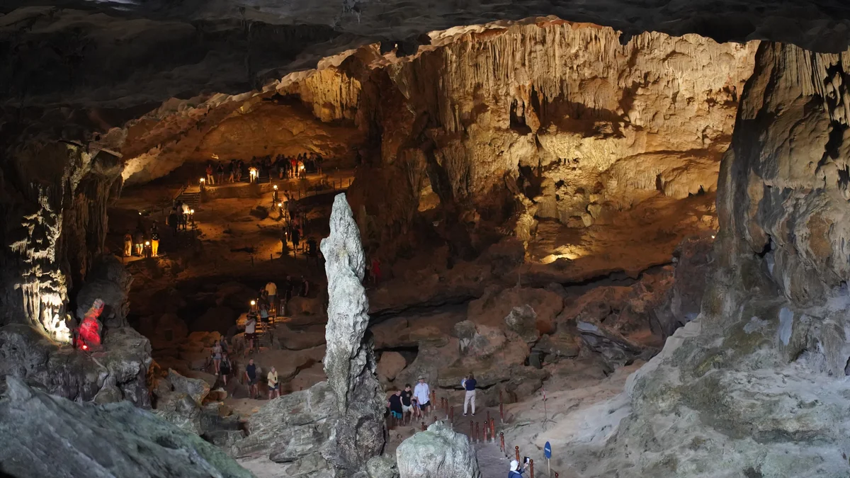 People exploring a large, illuminated cave with stalactites and stalagmites. Warm lighting highlights rocky textures. Dim, mysterious atmosphere.