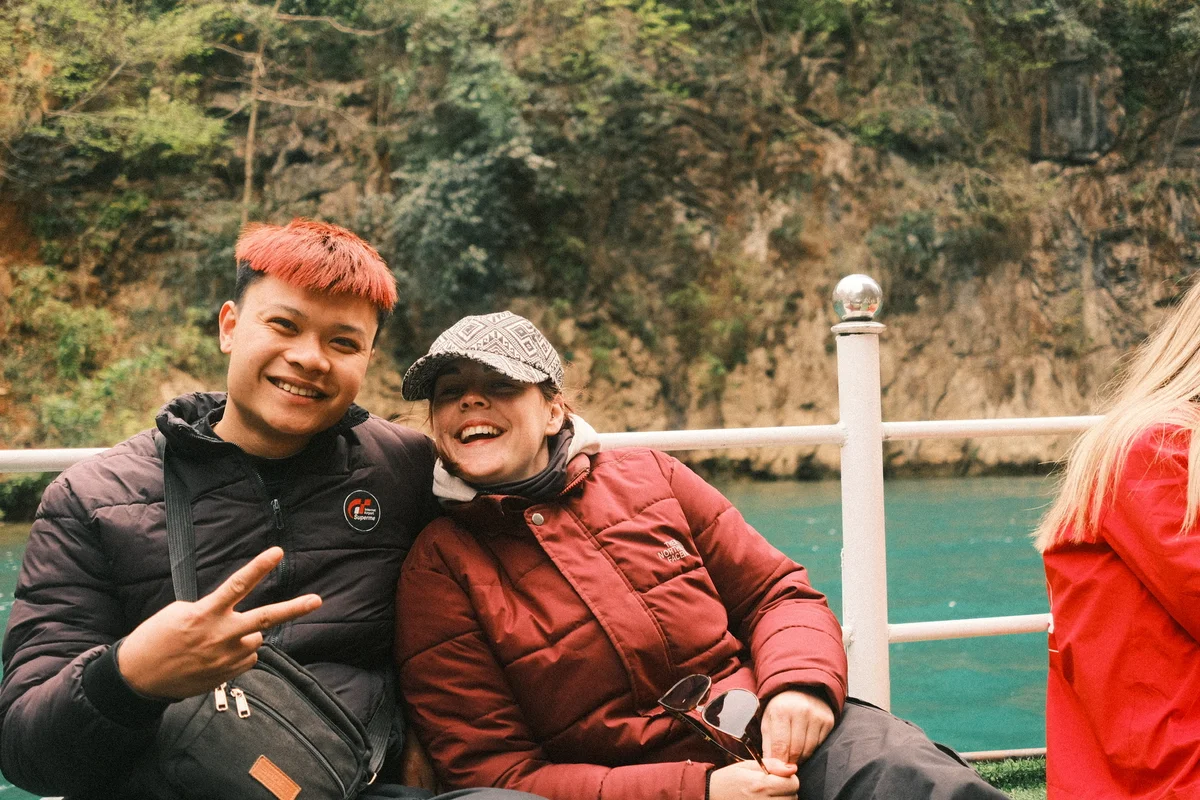 Two people in jackets smiling on a boat, with a rocky, forested background. One shows a peace sign; mood is cheerful. Water is visible.