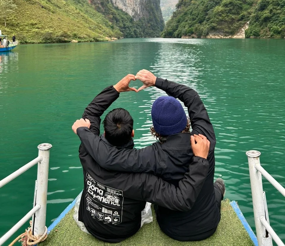 Two people sit on a boat, creating a heart shape with hands, overlooking a tranquil green river between lush hills, wearing dark jackets.