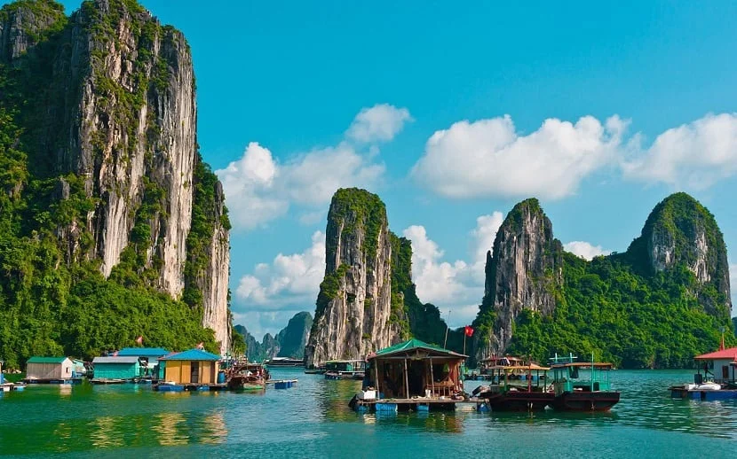 Boats float on turquoise water with towering green limestone cliffs in the background. Clear blue sky with fluffy clouds creates a serene mood.