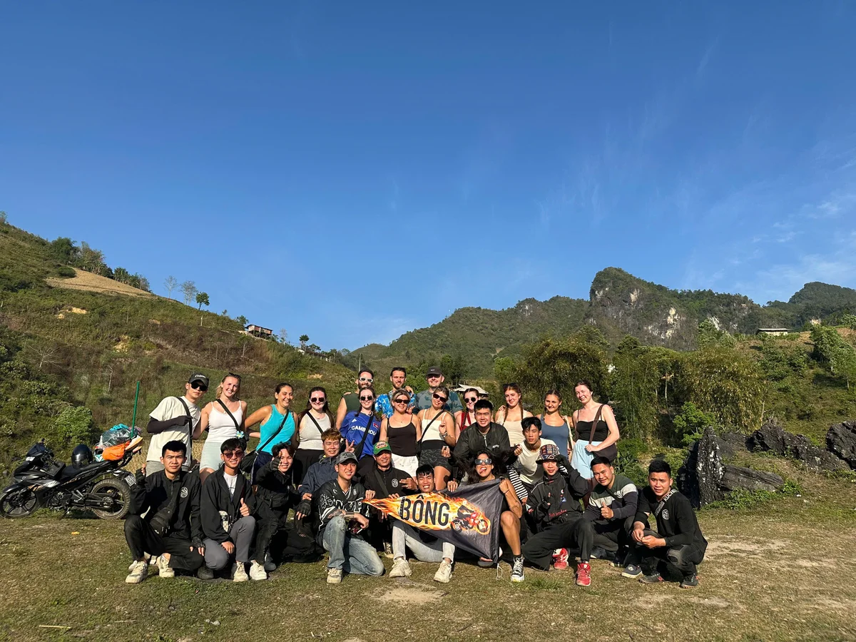 Group of people posing with a "Bong" banner in a hilly landscape. Clear blue sky, greenery, and motorcycles in the background. Everyone is smiling.