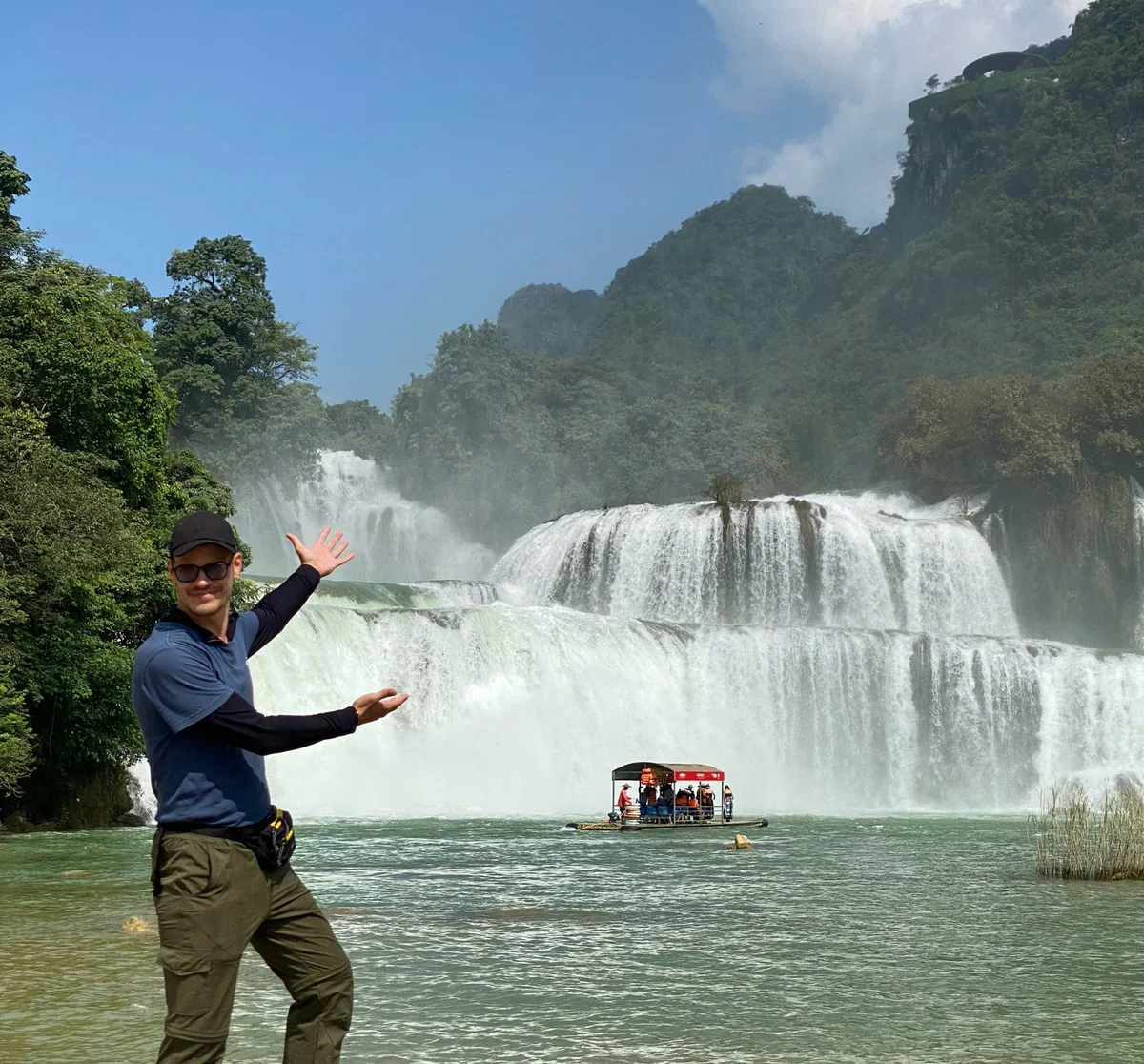 A person in sunglasses gestures at a scenic waterfall with a boat on the water. Green hills and a clear blue sky form the backdrop.