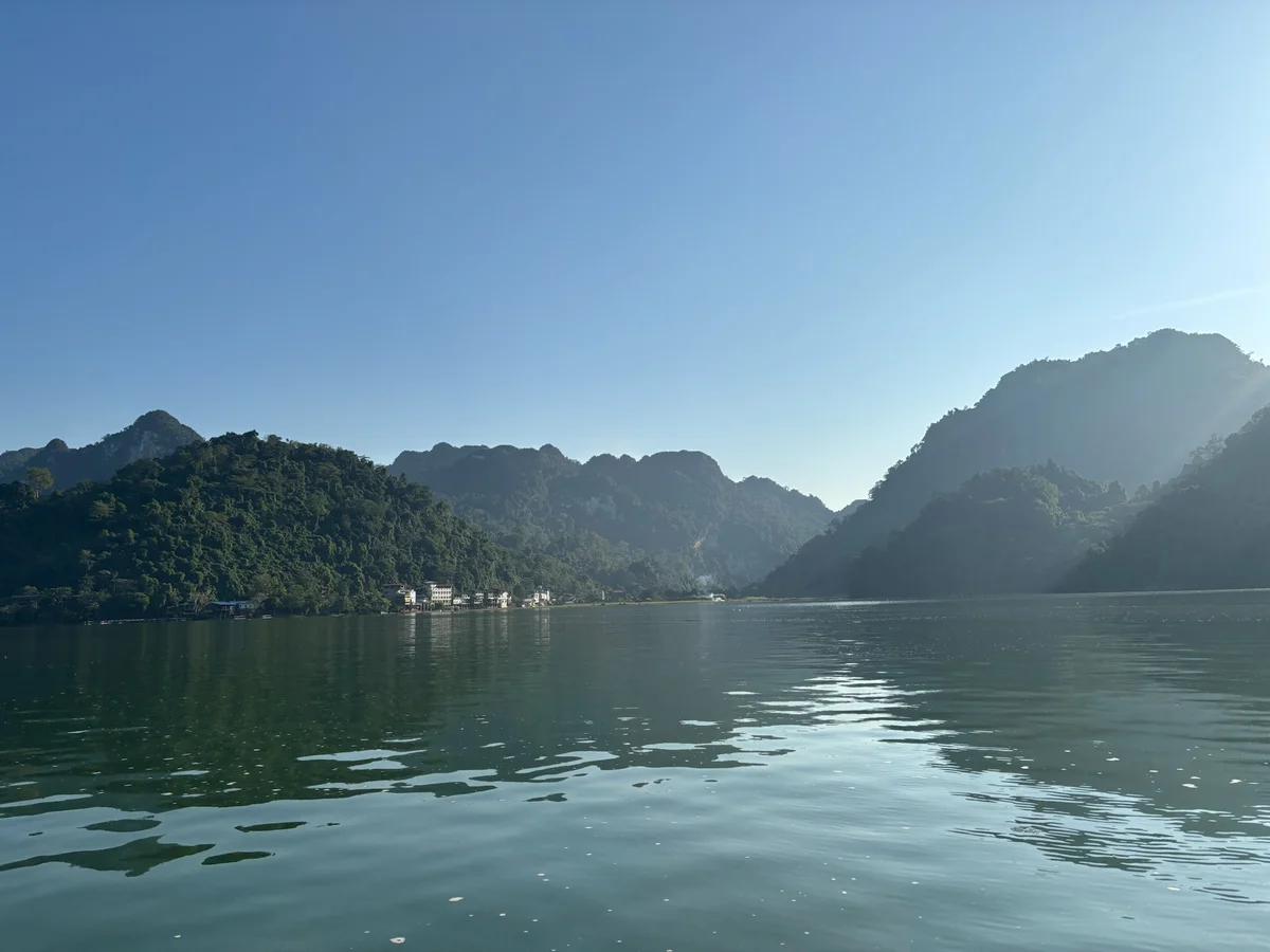 Calm lake reflecting lush green hills under a clear blue sky. Small buildings and gentle sunlight on the hillsides create a serene scene.