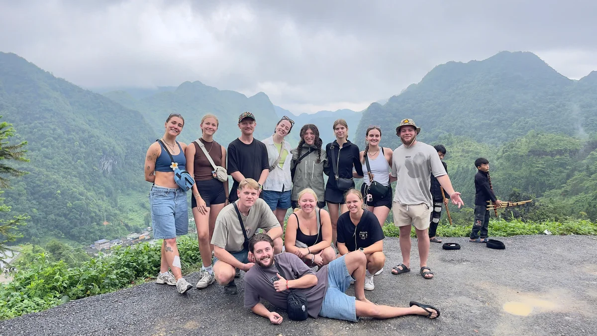Group of smiling people posing outdoors with misty green mountains in the background. Overcast sky, casual clothing, relaxed mood.