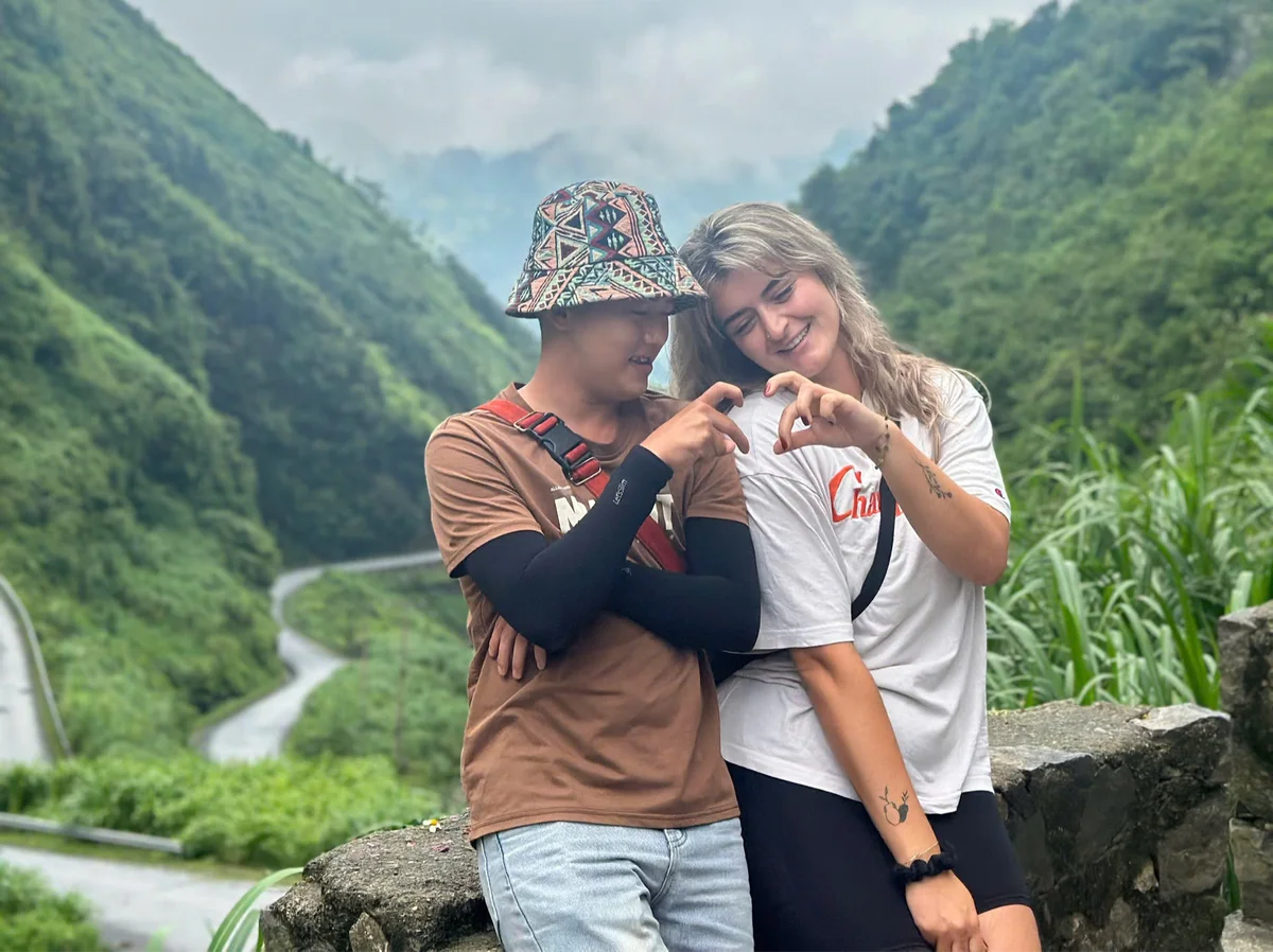 Couple forms a heart shape with fingers, smiling in lush green mountain setting. Road winds through the background. Mood is joyful.