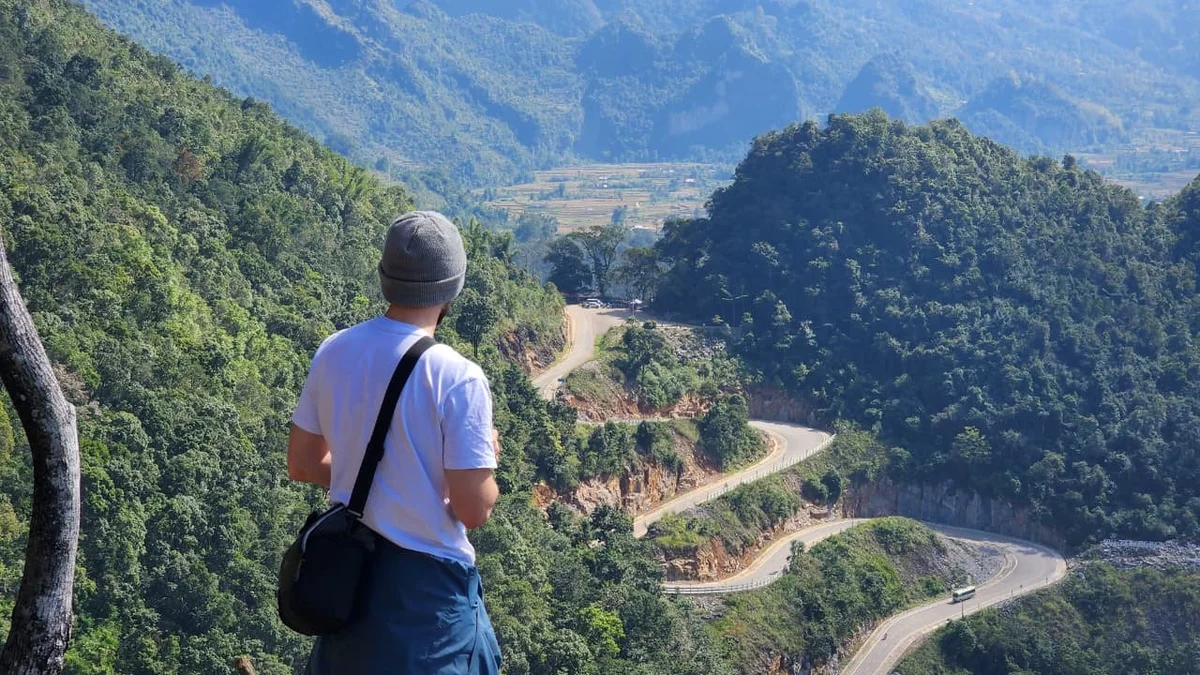 Man in a white shirt and beanie overlooks a winding mountain road, surrounded by lush greenery and distant hills under a clear sky.