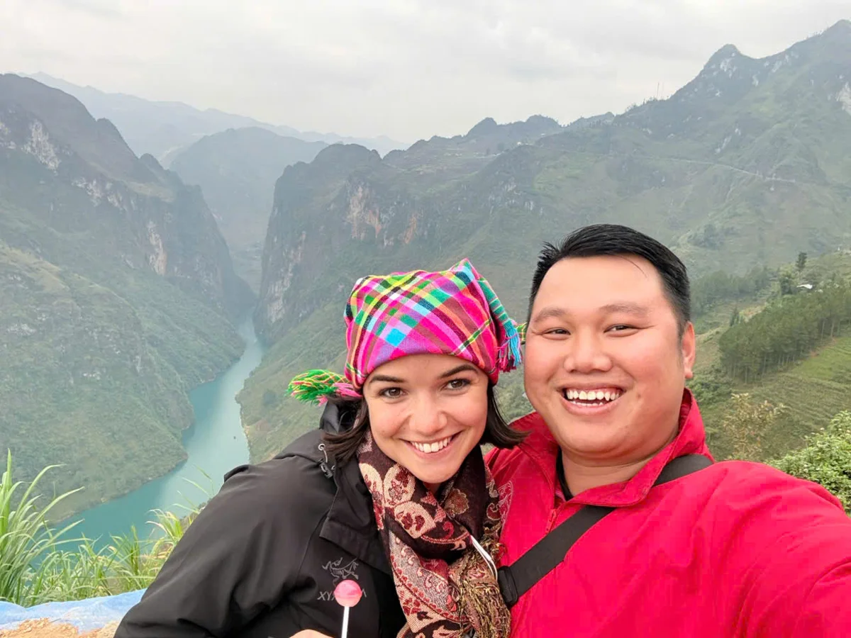Two smiling people taking a selfie overlooking a lush valley with a winding river. The woman wears a colorful headscarf, and both appear joyful.