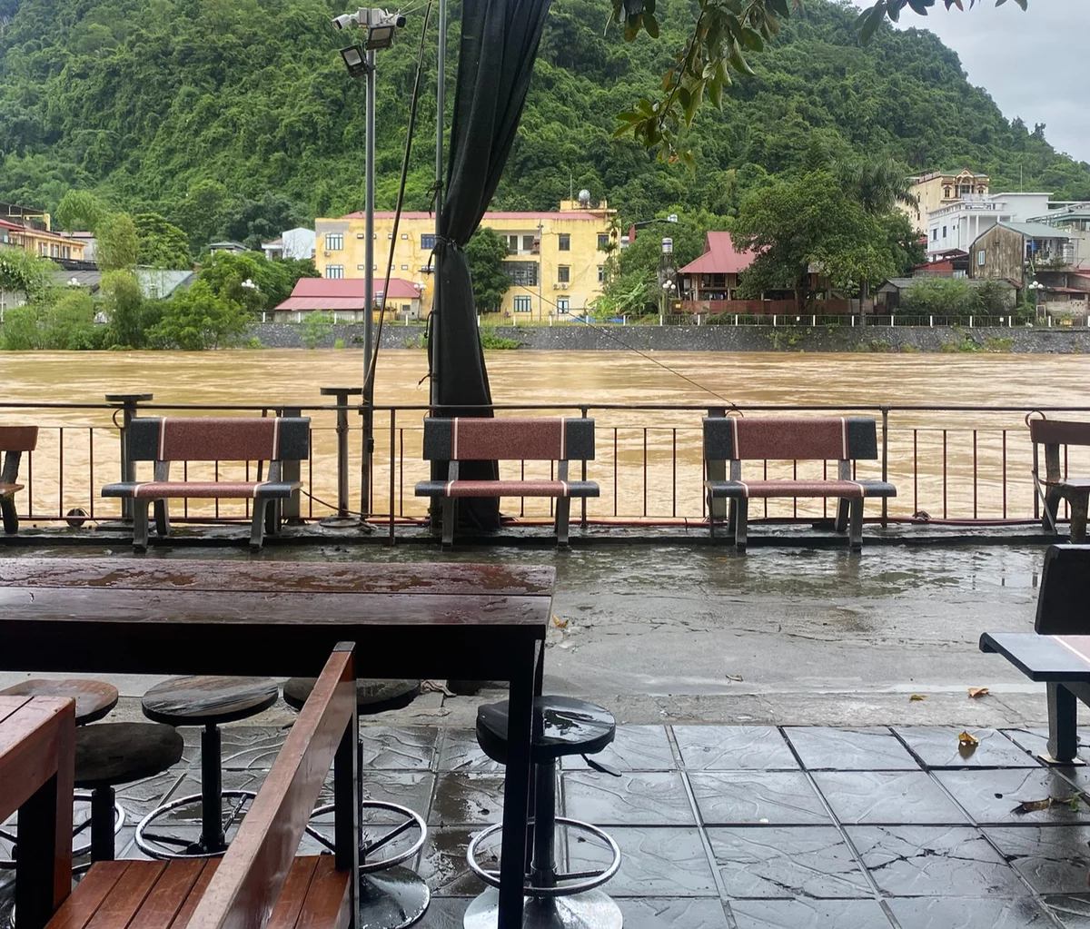 Empty riverside seating with wet benches, brown murky river, lush green hills, and colorful buildings in the background. Overcast mood.