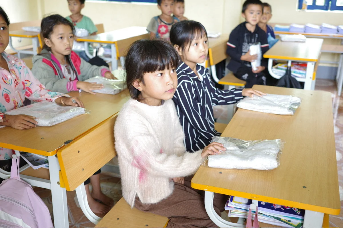 Children seated at wooden desks in a classroom, holding packages. The setting is bright, with books on shelves. The mood is attentive.