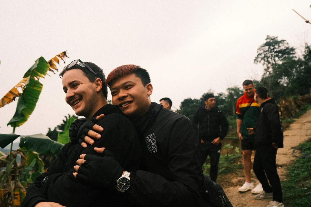 Two people embrace and smile in a rural setting, with others chatting in the background. Greenery and a dirt path are visible.