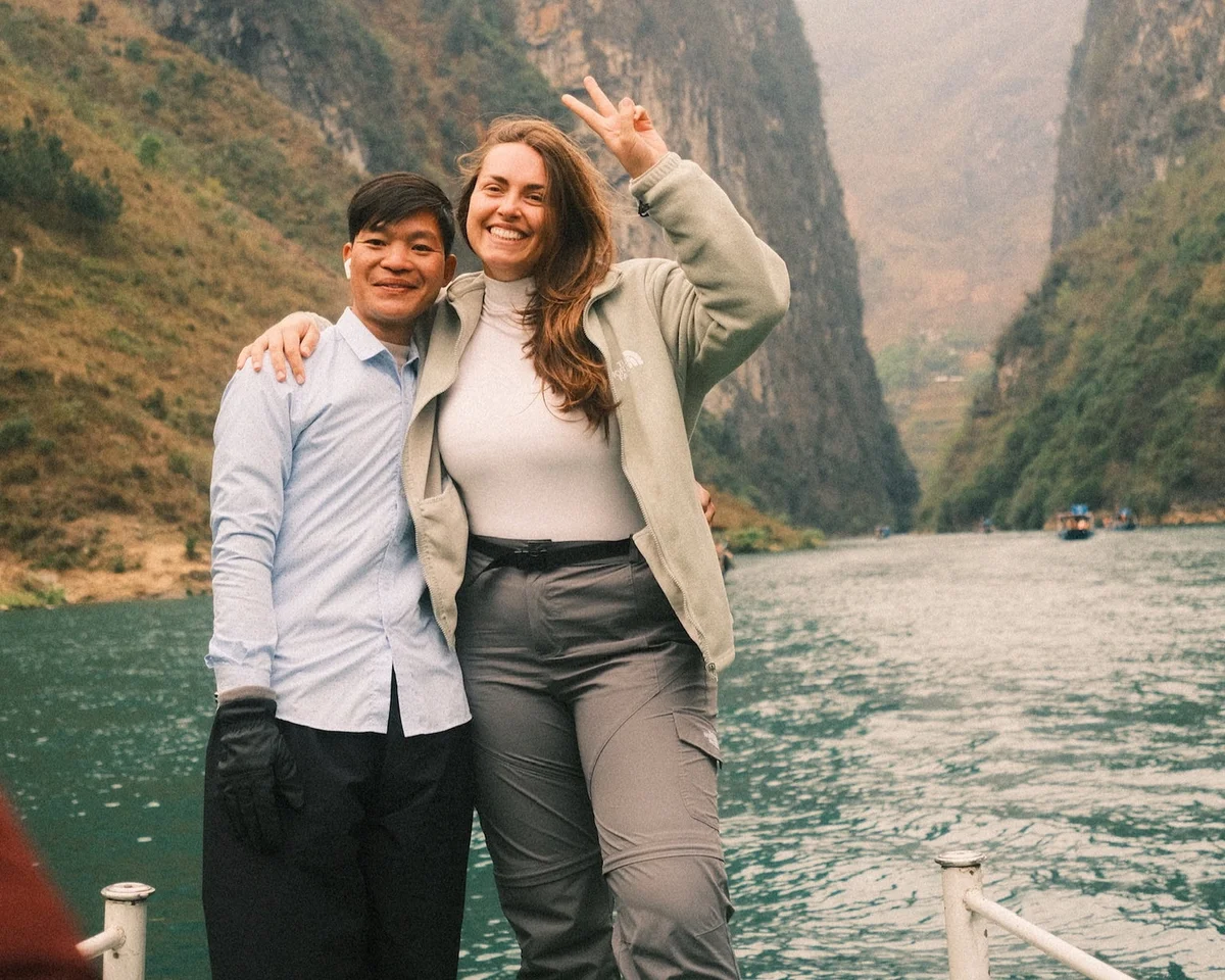 Two people smiling on a boat in a scenic river canyon. One flashes a peace sign. Misty cliffs in background, creating a happy, adventurous mood.