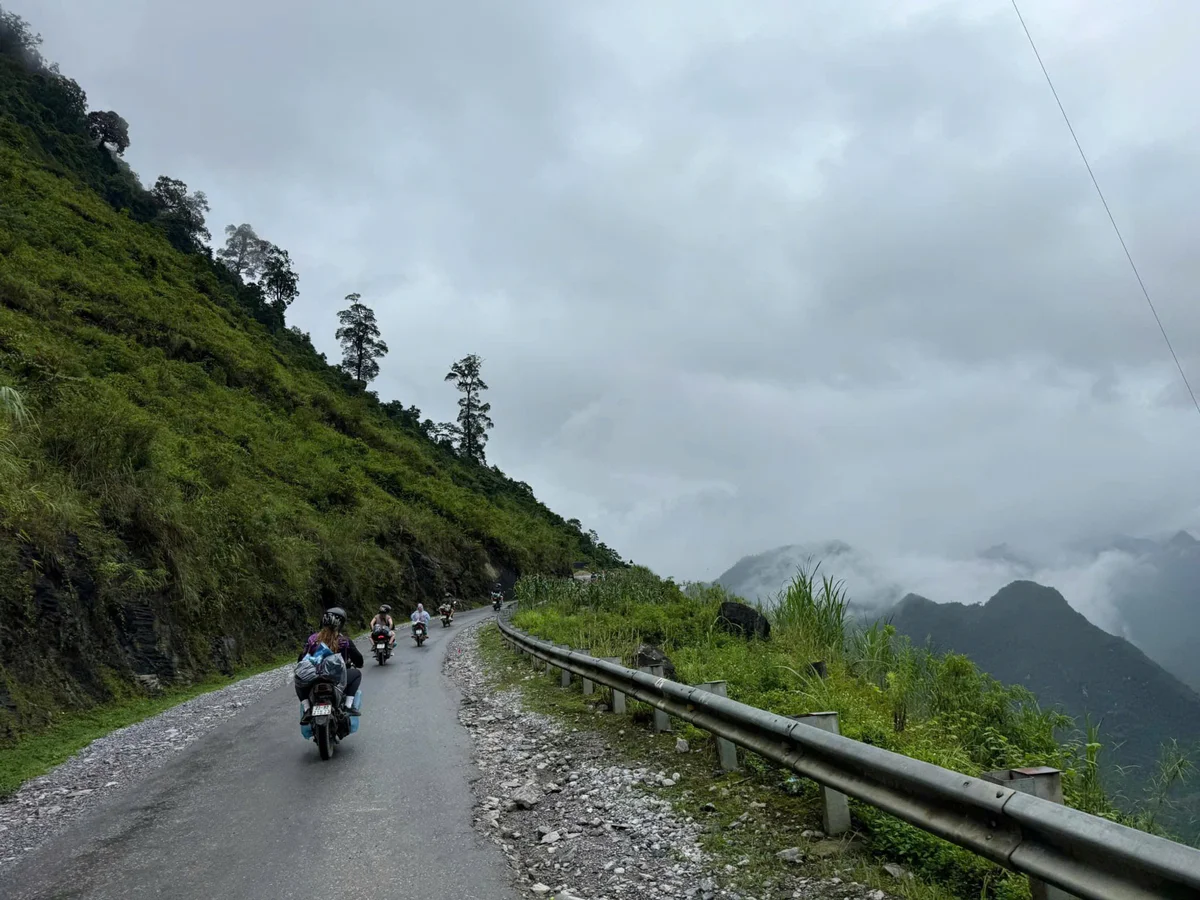 Motorcyclists ride on a narrow, misty mountain road bordered by lush greenery. Overcast sky and clouds create a serene, adventurous mood.