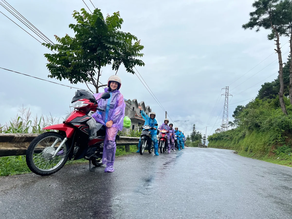 People in colorful raincoats stand with motorbikes on a wet, rural road. Greenery and trees line the background under a gray sky.
