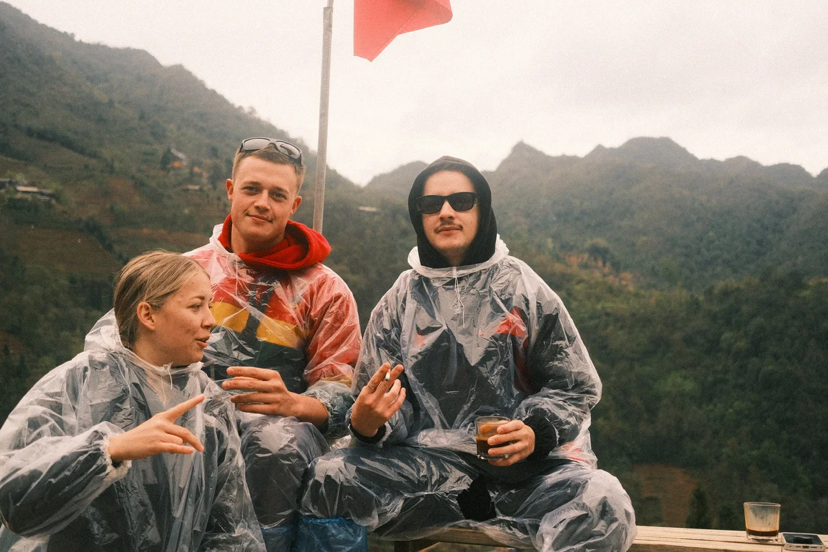 Three people in rain ponchos relax outdoors with drinks, against a backdrop of misty mountains. A red flag waves behind them.