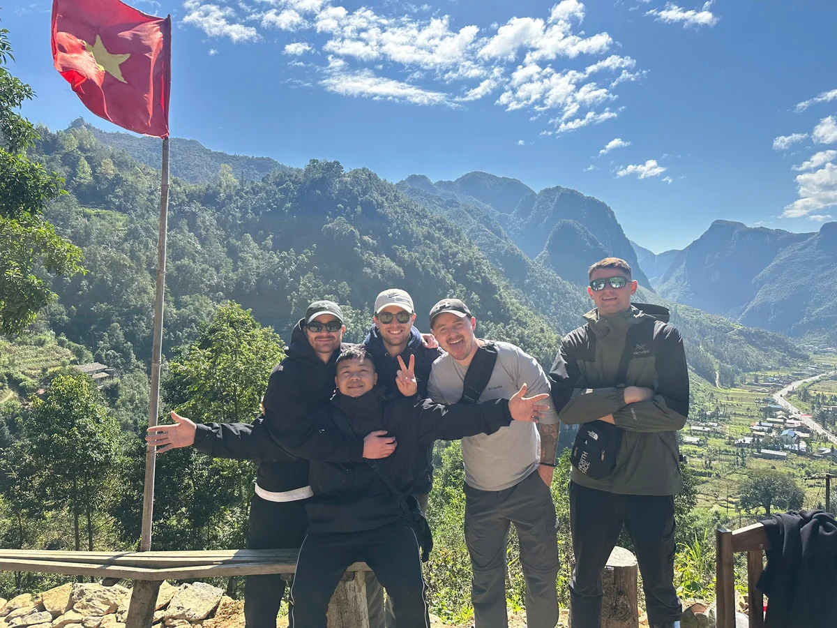 Five people pose joyfully on a bench in front of mountainous scenery under a blue sky. A red flag with a star waves on the left.
