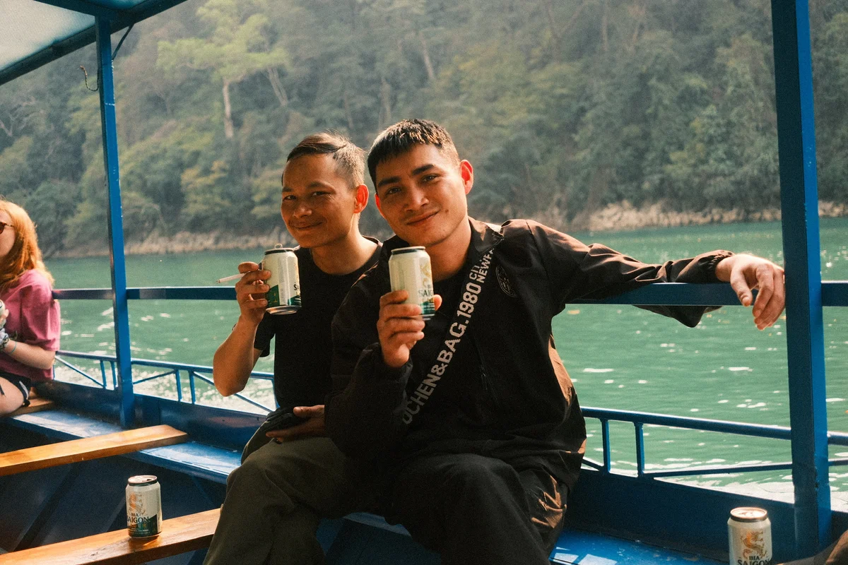 Two people smiling, holding cans on a blue boat, with a forest and lake in the background. A woman in pink is sitting nearby. Relaxed mood.