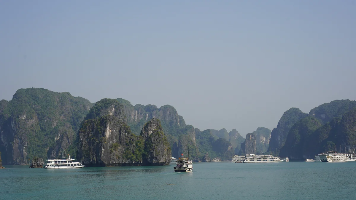Boats on tranquil turquoise water surrounded by towering green limestone cliffs under a clear blue sky. Peaceful and scenic atmosphere.