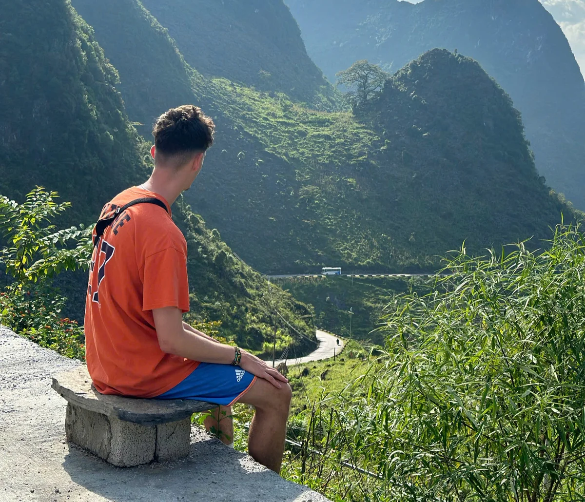 A man in an orange shirt and blue shorts sits on a stone bench, overlooking a lush green mountain road with a distant bus.