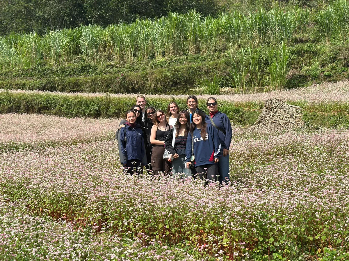 A group of people smiling in a lush field of pink flowers. Green plants and trees form a scenic backdrop under a sunny sky.