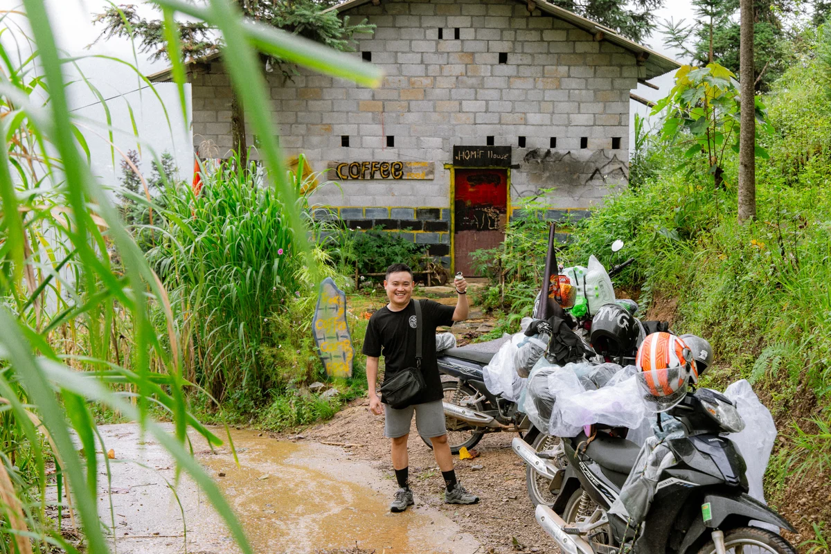 Man smiling beside motorbikes on muddy path, lush greenery and a rustic building labeled "Coffee" in the background, vibrant helmets visible.