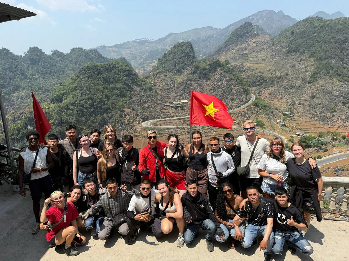 A group poses with red flags on a sunny mountain overlook. Winding road and green hills are visible in the background. Casual, joyful mood.