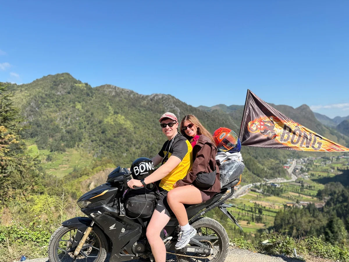 Two people on a black motorcycle with "BONG" helmets and flag. They're on a scenic mountain road under a clear blue sky, smiling.