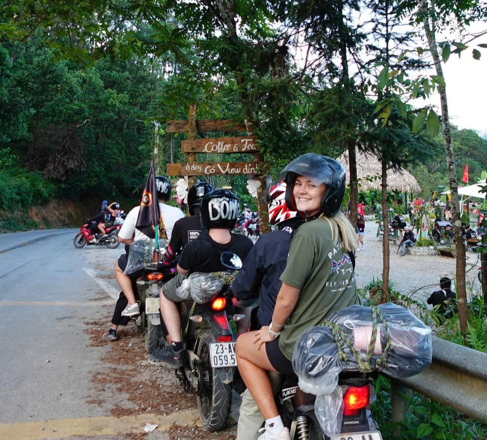 Motorcyclists parked by a road surrounded by greenery. A woman in a helmet smiles at the camera. A sign reads Coffee & Tea. Lively atmosphere.