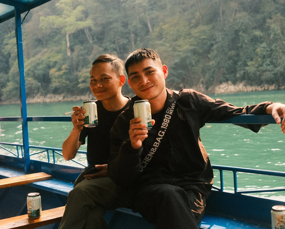 Two men smiling, seated on a boat, holding cans of Bia Saigon. One is smoking. Green water and forested background create a relaxed vibe.