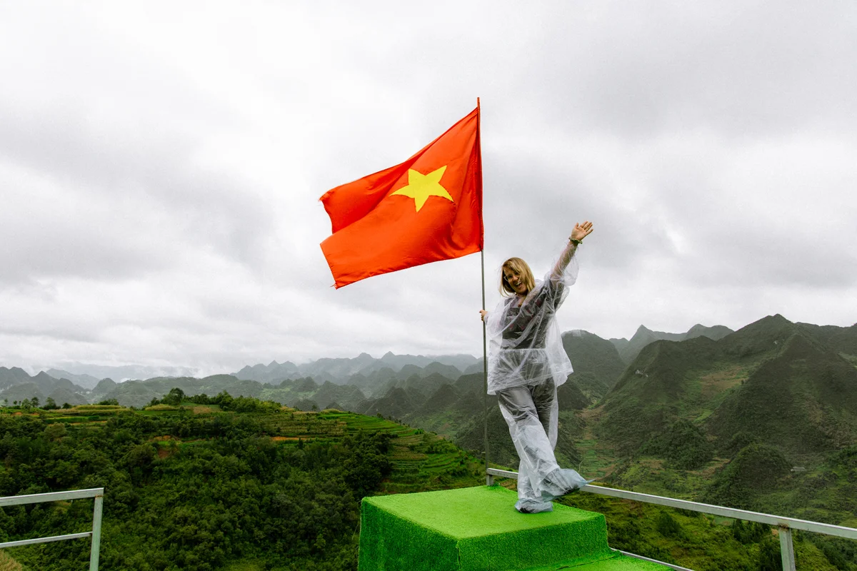 Woman in raincoat joyfully waving beside a large red Vietnamese flag on a green platform. Lush mountains under cloudy skies in background.