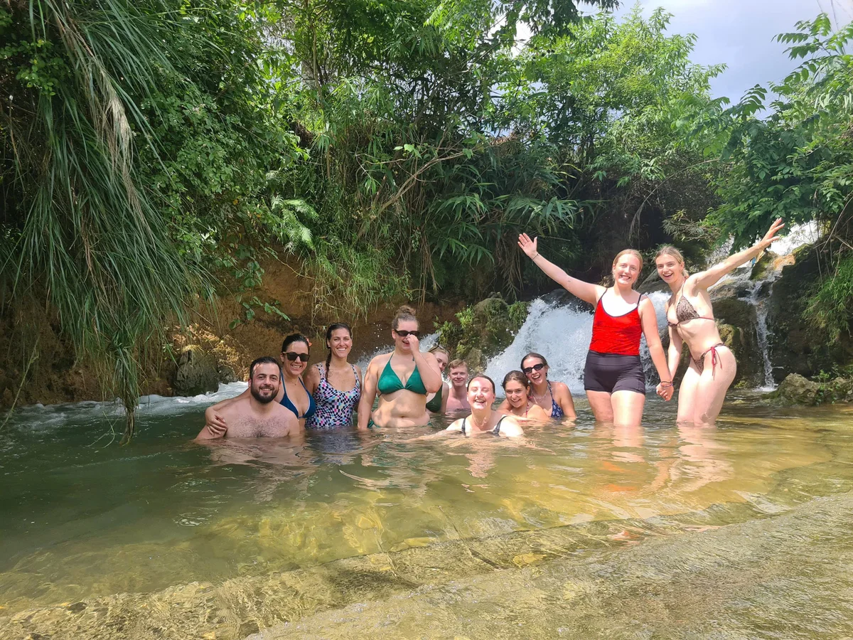 Group of people smiling and posing in a natural pool with a waterfall background. They're wearing swimsuits, surrounded by lush greenery.