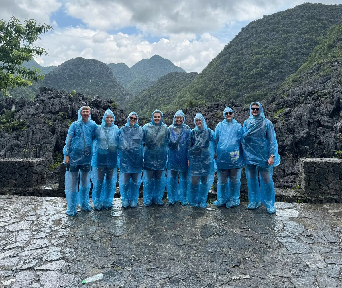 Group of people in blue rain ponchos stand in front of rocky mountains and cloudy sky, set on a wet stone path. Mood is cheerful yet damp.
