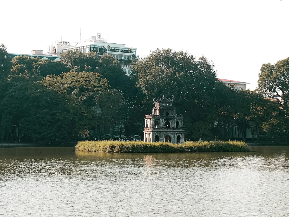 A historic stone tower on a grassy island in a tranquil lake, surrounded by lush trees. Buildings peek above the greenery in the background.