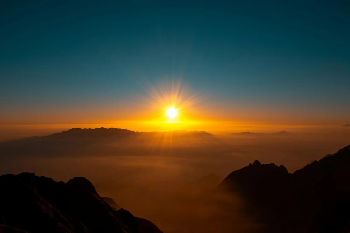 Sunrise over misty mountains, casting orange and yellow hues in the sky. Silhouetted peaks create a serene, tranquil atmosphere.