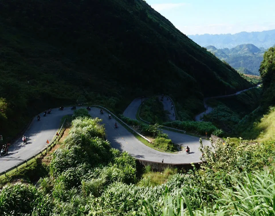 Motorcyclists navigate a winding mountain road surrounded by lush greenery and hills under a clear blue sky, creating a serene vibe.
