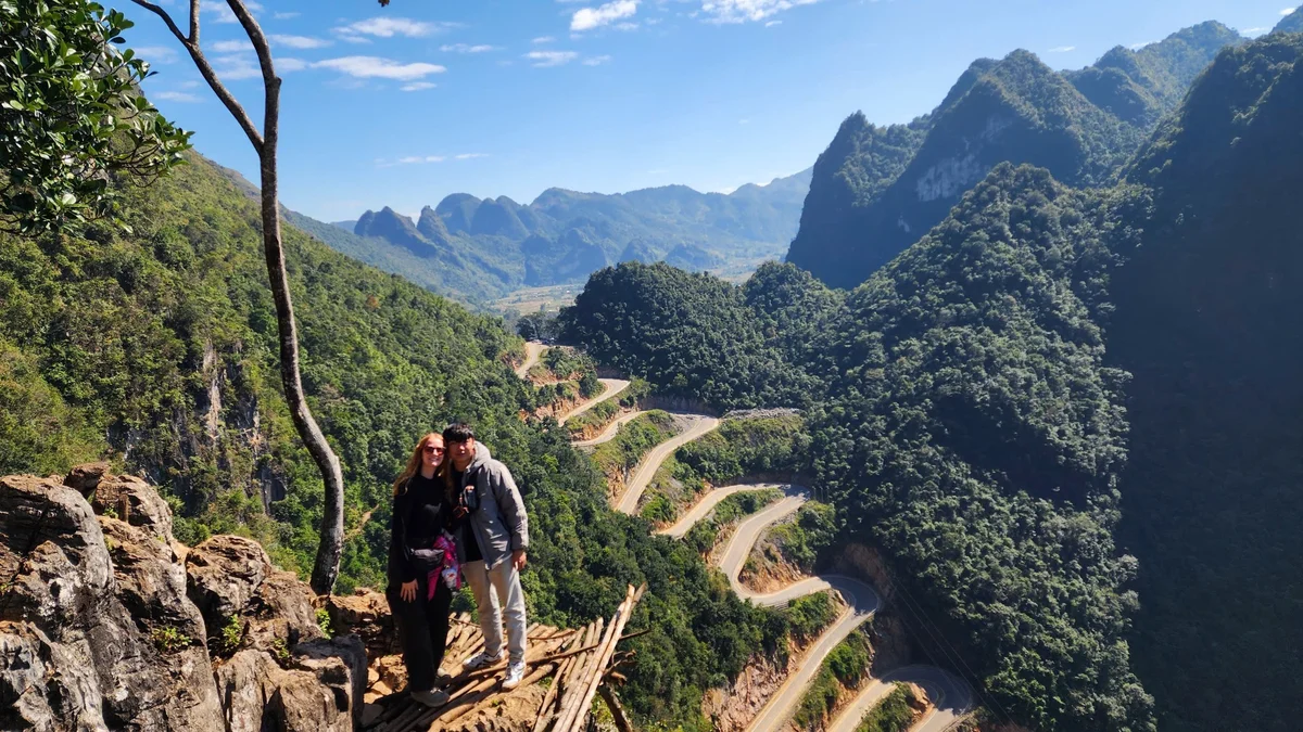 A couple stands on a rocky ledge overlooking a lush, winding mountain road under a clear blue sky, conveying a sense of adventure.