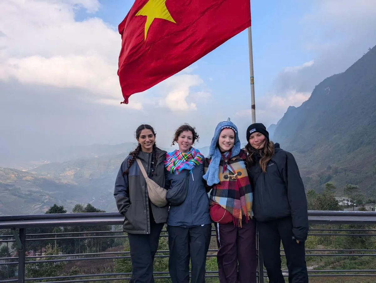 Four people in winter clothing smile under a waving red flag with a yellow star, against a mountainous backdrop and cloudy sky.