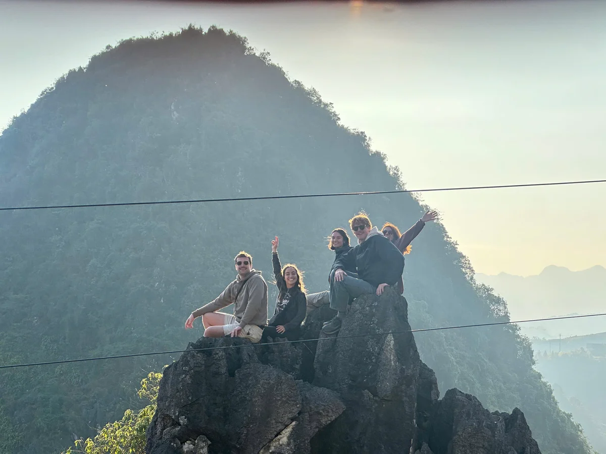 Group of five people sitting on rocks, smiling and waving, with misty mountains and a hazy sky in the background. Scenic and joyful mood.