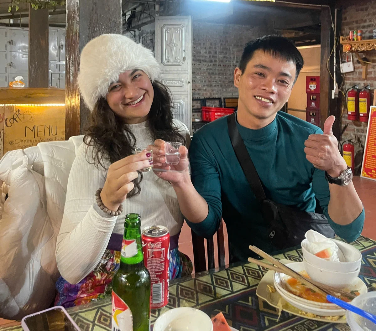Two smiling people holding drinks at a patterned table in a cozy restaurant. A woman in a white hat and a man gives a thumbs-up; festive mood.