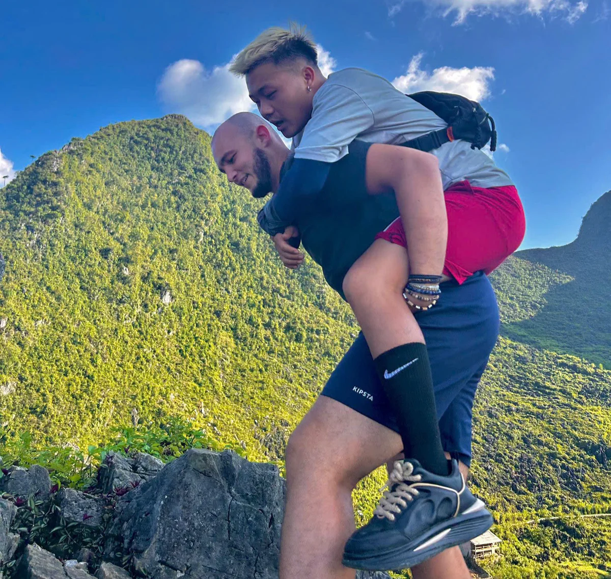Man gives another a piggyback ride on a rocky mountain trail, surrounded by lush green hills. Bright blue sky and sunny weather.