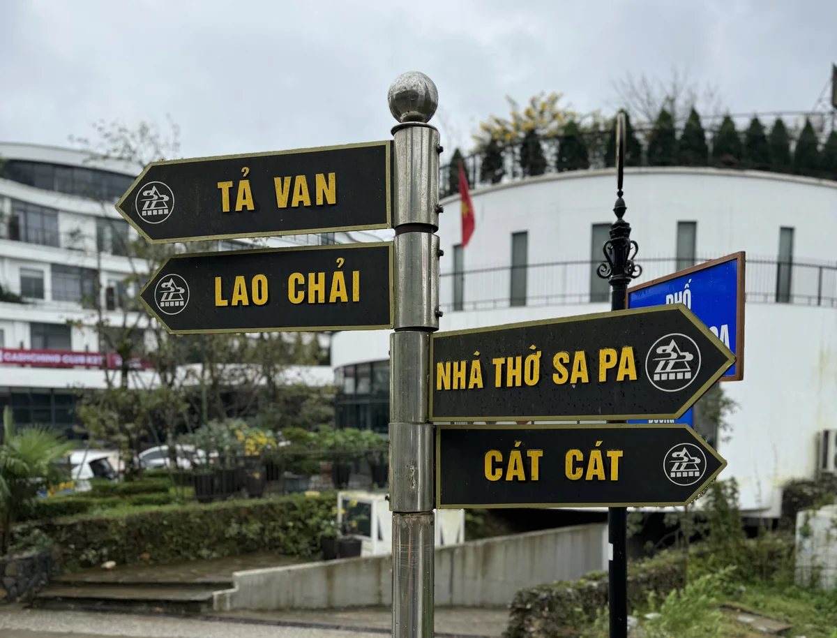 Street signpost with arrows to Tả Van, Lao Chải, Nhà Thờ Sa Pa, and Cát Cát. Overcast day, white building in the background.